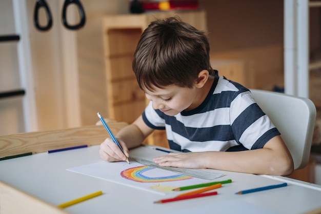 adorabile ragazzo caucasico di età scolare che disegna un arcobaleno con le matite seduto alla scrivania