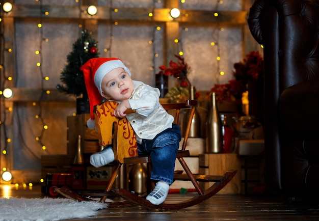 Adorabile ragazzo con il cappello rosso di Babbo Natale sorride e tiene un cavallo a dondolo sullo sfondo delle luci festive