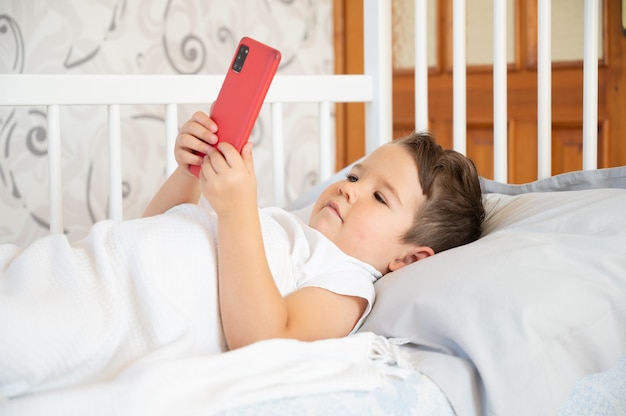 Adorabile ragazzo di 3 anni che guarda lo smartphone in camera da letto. Foto di alta qualità