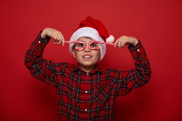 Adorabile ragazzo in cappello di Babbo Natale e camicia a quadri guarda la telecamera attraverso dolci lecca-lecca, tenendoli imitando gli occhiali, posando su uno sfondo colorato con spazio copia per la pubblicità di Natale