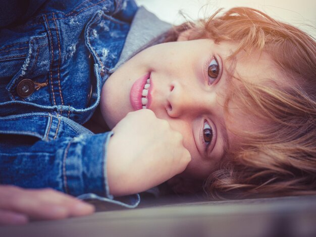 Adorabile ragazzo in giacca di jeans con capelli biondi e occhi castani che tocca il viso e guarda la fotocamera mentre giace a terra