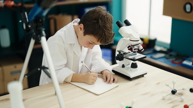 Adorabile ragazzo ispanico che usa la scrittura al microscopio sul taccuino in classe di laboratorio