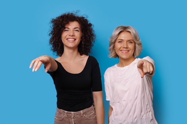 Adorabili donne in abiti casual stanno indicando la telecamera sorridendo su una parete blu con spazio libero