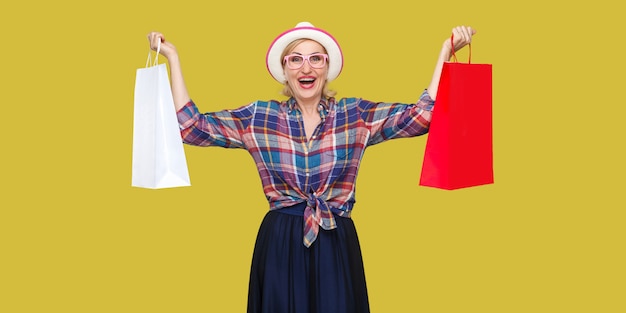 Adoro lo shopping! Soddisfatta moderna nonna con cappello bianco e camicia a scacchi che tiene in mano borse della spesa e trionfa con le braccia alzate e un sorriso a trentadue denti. Indoor, studio shot isolato su sfondo giallo.