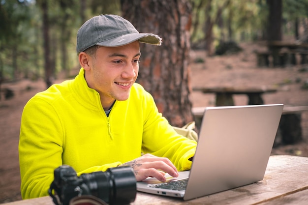 Affari all'aria aperta per un bellissimo giovane libero professionista che lavora su un computer portatile seduto all'aperto nella foresta Viaggiatore ragazzo hipster che lavora a distanza mentre si gode la montagna della natura durante le vacanze