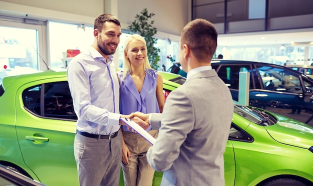 affari automobilistici, vendita di automobili e concetto di persone - coppia felice con concessionario che stringe la mano in un salone di auto o in un salone