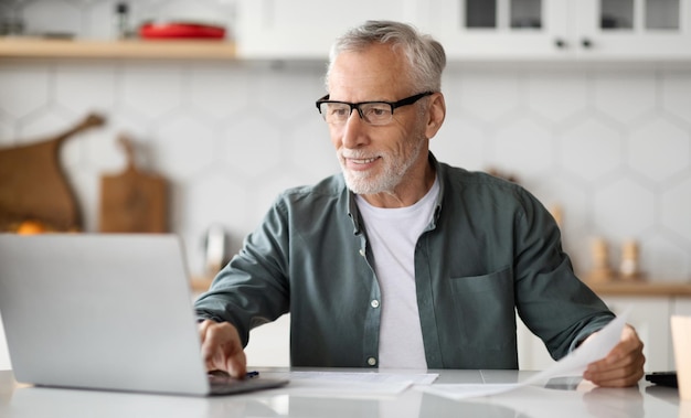 Affari dopo il pensionamento uomo anziano sorridente che lavora con il computer portatile nell'interno della cucina