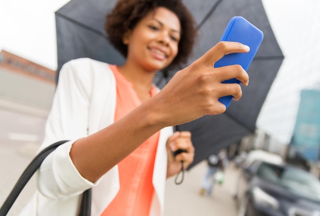 affari, maltempo e persone e concetto - primo piano di felice donna d'affari afroamericana con ombrello che manda messaggi sullo smartphone in una strada cittadina