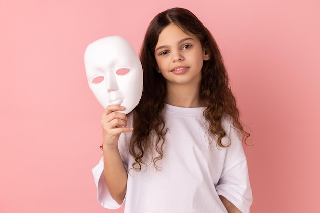 Affascinante adorabile bambina dai capelli scuri che tiene in mano una maschera bianca vuole cambiare personalità