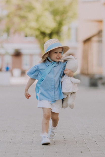 affascinante bambina bionda con un cappello di paglia e un vestito blu gioca con un coniglio farcito all'aperto