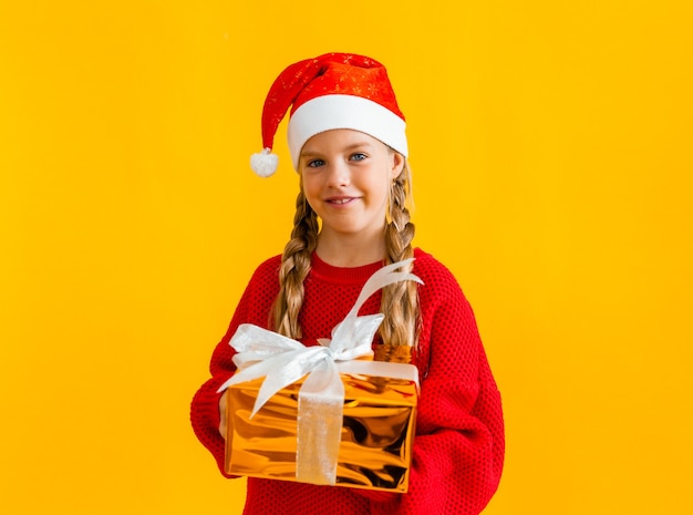 Affascinante bambina con un maglione lavorato a maglia e un cappello da Babbo Natale con un regalo su uno sfondo giallo.