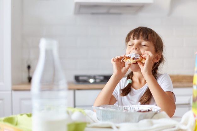 Affascinante bambina dai capelli scuri affamata che morde e si gode il croissant, tenendo gli occhi chiusi, seduta al tavolo in cucina, assaggiando la deliziosa pasticceria fatta dalla sua adorabile mamma.