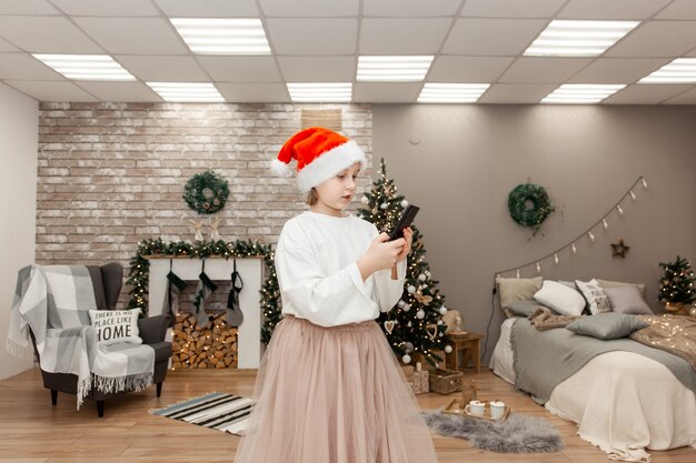 Affascinante bambina di bell'aspetto che parla al telefono con qualcuno, si congratula, ha due codini, indossa un cappello da Babbo Natale, guarda la telecamera mentre posa in soggiorno con decorazioni natalizie.