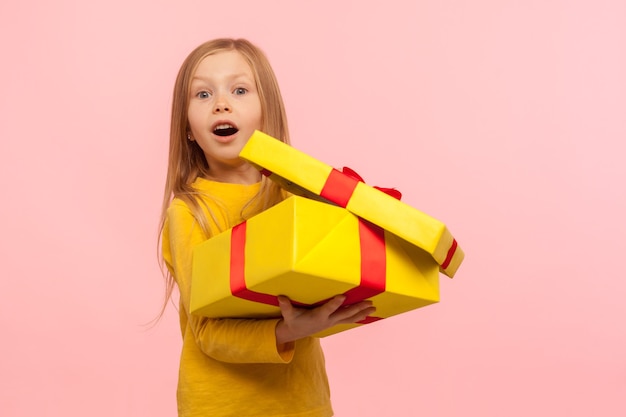 Affascinante bambino sorpreso dal regalo di compleanno. Ritratto di una bambina carina che apre una confezione regalo e tiene la bocca aperta con stupore, espressione scioccata. studio al coperto isolato su sfondo rosa