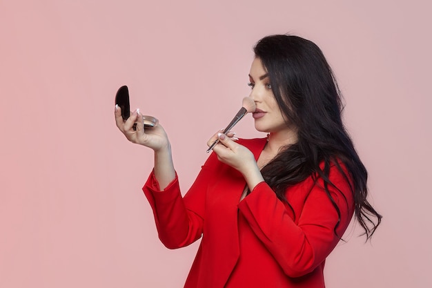 Affascinante bruna con i capelli lunghi tiene una compatta e un pennello tra le mani. servizio fotografico su sfondo rosa