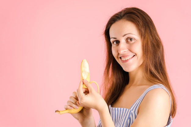 Affascinante bruna sorride e sbuccia una banana su uno sfondo rosa, copyspace