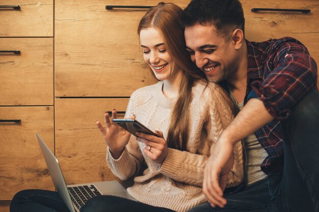 Affascinante coppia caucasica divertendosi al mattino sul pavimento della loro cucina guardando lo schermo di uno smartphone che tiene un computer portatile.