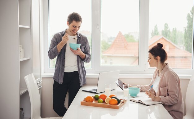 Affascinante coppia mangiare cereali con latte e fare lezioni online utilizzando tablet e laptop