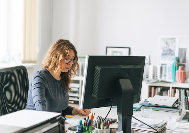 Affascinante donna adulta sorridente donna di mezza età con capelli ricci designer che lavora al computer in ufficio