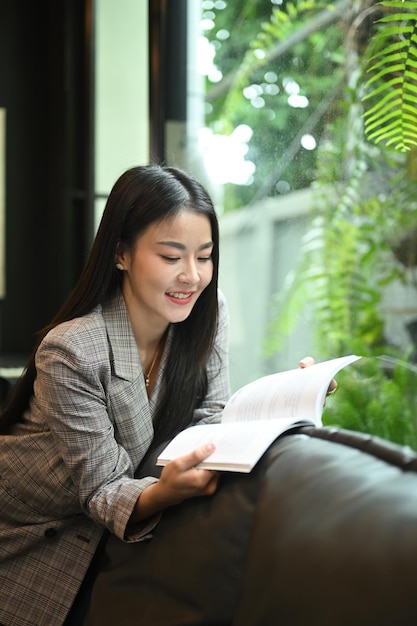 Affascinante donna asiatica che si prende una pausa dal lavoro e legge un libro sul comodo divano