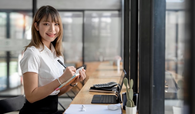 Affascinante donna asiatica con un sorriso che guarda l'obbiettivo mentre è seduto e tiene in mano il taccuino in ufficio