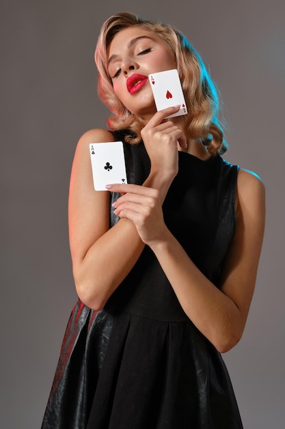 Affascinante donna bionda con trucco luminoso in abito nero elegante mostra due carte da gioco e posa su sfondo grigio studio Concetto di gioco d'azzardo intrattenimento poker casinò Primo piano