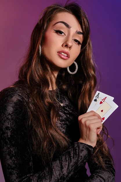 Affascinante donna bruna con trucco, in abito di velluto nero e gioielli sta mostrando due carte da gioco e ti sta guardando, in posa su sfondo colorato. Gioco d'azzardo, poker, casinò. Avvicinamento.