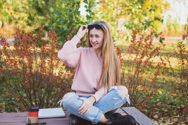 Affascinante donna con capelli lunghi e occhiali da sole casual si siede su una panchina nel bellissimo parco autunnale