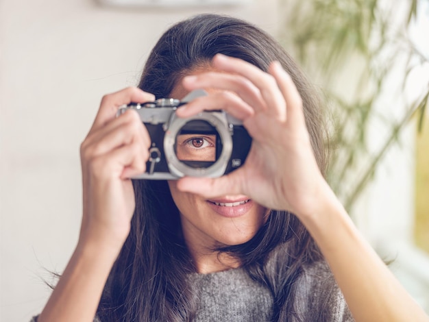 Affascinante donna con lunghi capelli scuri e occhi castani che guarda attraverso una macchina fotografica analogica retrò mentre finge di scattare foto e guarda la fotocamera su sfondo sfocato