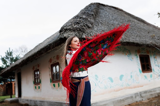 Affascinante donna con tradizionale collana fazzoletto ucraino e abito ricamato in piedi sullo sfondo di capanna decorata con tetto di paglia Cultura etnica popolare in stile ucraino