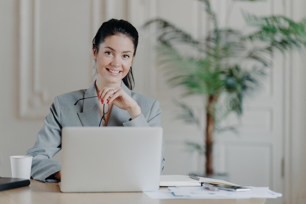 Affascinante donna d'affari elegante lavora come freelance al computer portatile utilizza la connessione Internet ad alta velocità tiene gli occhiali in mano gode del lavoro remoto preferito legge la pubblicazione si siede alla scrivania dell'ufficio