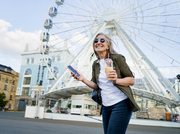 Affascinante donna europea dai capelli grigi felice di godersi il tempo libero dopo il lavoro o in viaggio tenendo il telefono mentre beve un succo in movimento utilizzando un bicchiere di plastica in città e la ruota panoramica sullo sfondo