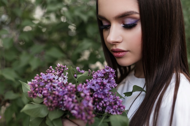 Affascinante donna in bandana in abiti bianchi con trucco con bouquet di incredibili fiori viola freschi si trova vicino a fogliame verde nel parco. La ragazza carina guarda il bouquet e gode di bellezza e odore di lillà.