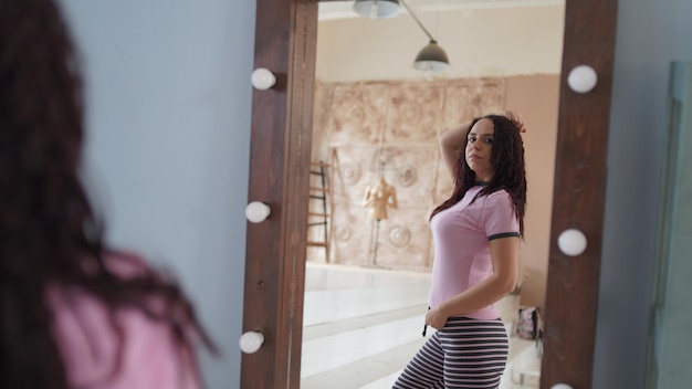 Affascinante donna in pigiama che si guarda allo specchio Vista laterale della giovane donna in pigiama viola che sorride mentre si guarda sopra la spalla nello specchio nella camera da letto moderna