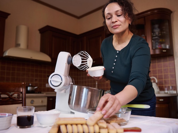 Affascinante donna multietnica pasticciera casalinga che prepara il tradizionale dessert italiano Torta tiramisù con biscotti biscotto savoiardi nella cucina di casa