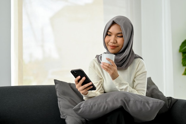 Affascinante donna musulmana asiatica in hijab che sorseggia caffè mentre usa il suo telefono cellulare sul divano