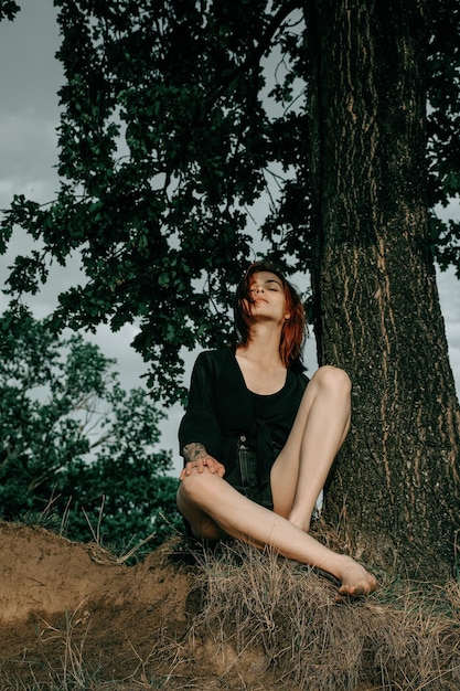 Affascinante donna rossa sexy con le gambe lunghe che si rilassano vicino a un albero con energia femminile.