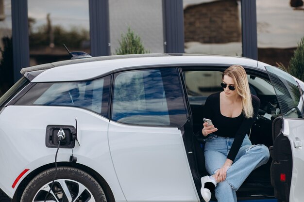 Affascinante donna seduta all'interno della ricarica di un'auto elettrica con uno smartphone moderno e una tazza di caffè in mano Femmina caucasica che parla tramite videochiamata mentre aspetta che la sua auto venga caricata