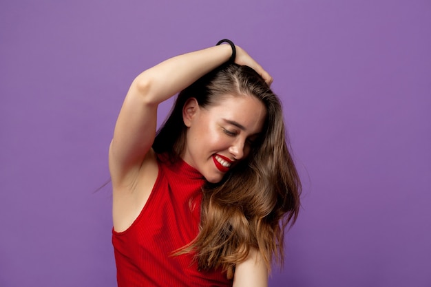 Affascinante donna sorridente con capelli ondulati che indossa top rosso sorridente con gli occhi chiusi e toccando i suoi capelli sulla viola
