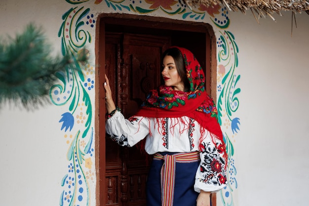 Affascinante donna sorridente in tradizionale collana fazzoletto ucraino e abito ricamato in piedi sullo sfondo della capanna decorata cultura etnica popolare in stile ucraino