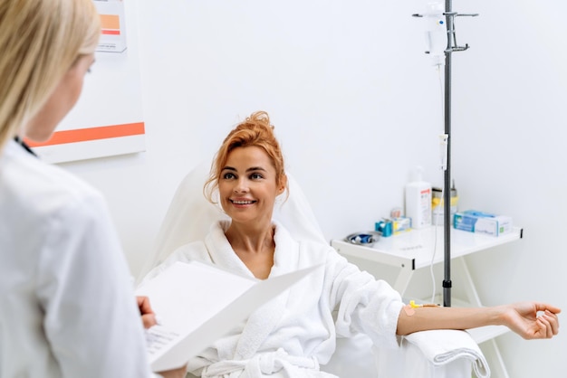 Affascinante donna zenzero in accappatoio bianco sdraiata sul lettino e guardando l'estetista con un sorriso mentre parla con lei e tiene la cartella