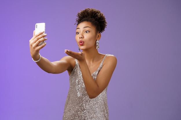 Affascinante ed elegante giovane studentessa che fa festa godendosi il ballo di fine anno registrazione video smartphone guarda il display del telefono invia un bacio d'aria che soffia mwah prendendo selfie tenere gadget, in piedi sfondo blu.