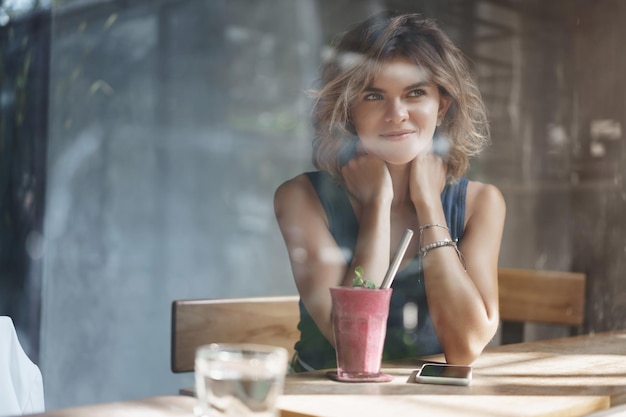 Affascinante giovane bionda dai capelli ricci ragazza impertinente sorridente sguardo sognante fuori dalla finestra deliziato tocco spalle flirty adorabile ghignante ricordando bel ricordo bere frullato da solo caffè