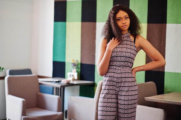 Affascinante giovane donna afroamericana elegante con lunghi capelli ricci che indossa una tuta in posa al bar al coperto