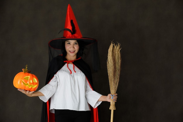 Affascinante giovane donna asiatica sorridente in costume da strega con zucca di Halloween e scopa guardando la telecamera su sfondo grigio scuro