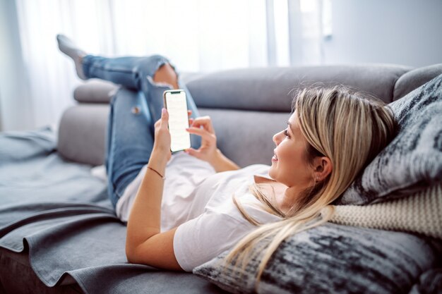 Affascinante giovane donna bionda caucasica vestita casual sdraiato sul divano nel soggiorno, rilassarsi e utilizzando smart phone.