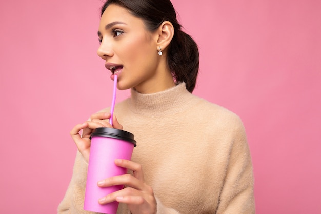 Affascinante giovane donna bruna felice che indossa abiti eleganti isolati su sfondo colorato parete tenendo il bicchiere di carta per mockup bere frullato guardando al lato