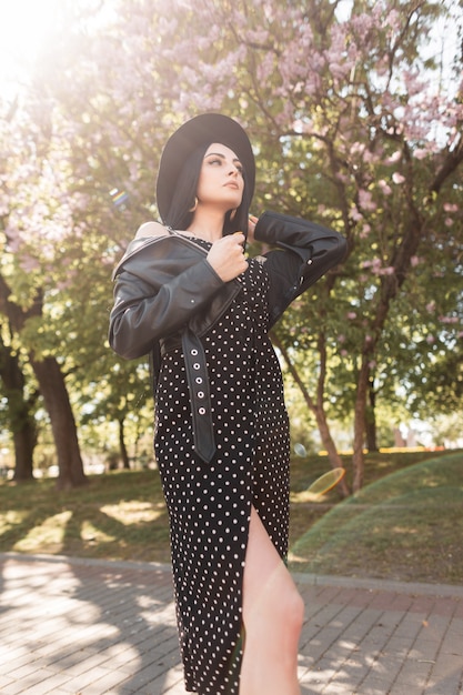 Affascinante giovane donna bruna in abito femminile estivo alla moda gode del sole e raddrizza il cappello elegante nel parco fiorito in una giornata di sole primaverile. La ragazza attraente in vestiti neri alla moda gode sulla natura.