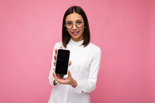 Affascinante giovane donna bruna sorridente che indossa camicetta bianca e occhiali ottici in piedi isolato su sfondo rosa che mostra il telefono cellulare con schermo vuoto per mockup guardando la fotocamera