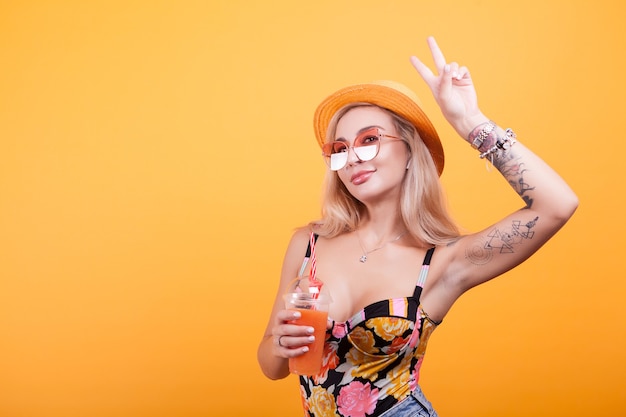 Affascinante giovane donna che fa segni di pace mentre si mangia un succo d'arancia fresco con occhiali da sole su bacground giallo in studio